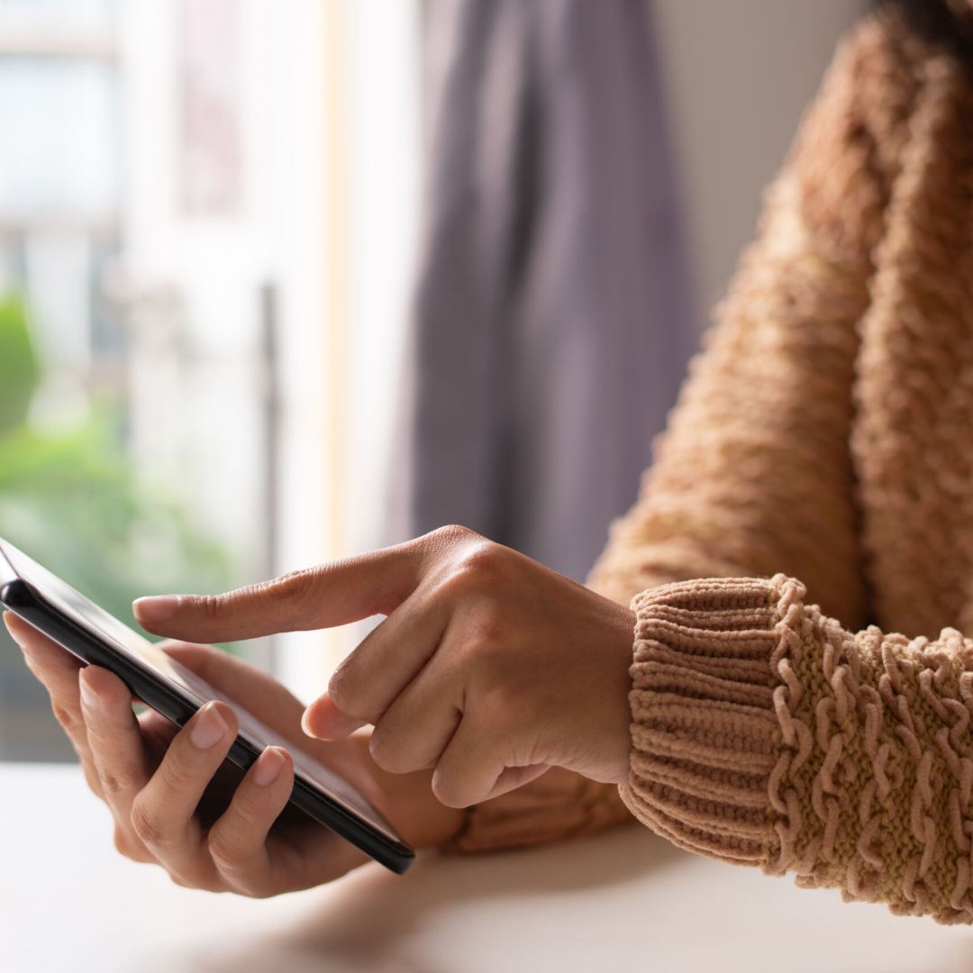 Close-up of girl in sweater viewing online information on smartphone. Unrecognizable lady sitting at table and using modern gadget. Technology concept
